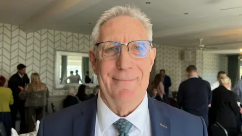 BBC Malcolm Lewis wearing a dark blue suit jacket, white shirt, and patterned blue tie stands in the foreground of a banquet-style room. Several groups of people are gathered and talking.