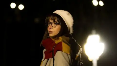 A woman walking home at night. She is wearing a coat and a hat and there is a street light behind her. She has a bag on her shoulder and she is wearing glasses and looking serious.