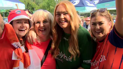 Four women celebrate. They're wearing orange and green gaa colours.