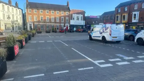 A market square with a combination of bold white lines market the road and more faded dotted lines where parking bays were once in place. To the left are planters with shrubs. To the right are parked vehicles. In the background are retail premises around three sides.