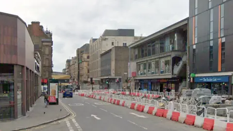 Google Retail and hospitality venues in buildings of mixed architectural styles line Cambridge Street, which has roadwork bollards along the middle of the road. 