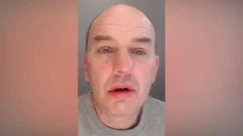 Neil Cook. He is bald and clean shaven and his eyes appear out of focus.