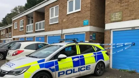 BBC A police vehicle parked outside a housing block. The block consists of a row of properties with two storeys. The living accommodation is on the second floor above garages, which have blue doors. Behind the police car there is police tape attached to a garage door, cordoning off an alley between two of the properties.