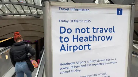 Reuters A travel information board with "Do not travel to Heathrow Airport" written in blue letters on it beside an escalator which has a woman in a black coat and blue jeans going down it