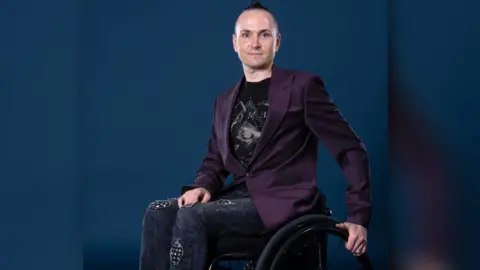 Olly Courtney Owen Tooth wears a purple blazer and looks directly at the camera. He has one hand on his lap and the other on the wheel of his wheelchair.