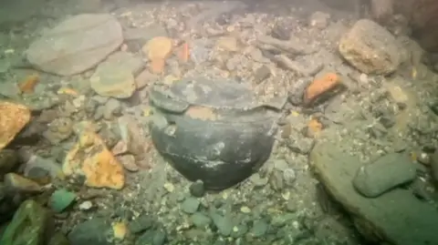 Mark Pearce A half exposed chamber pot which is rounded metal among rocks and shale underwater 