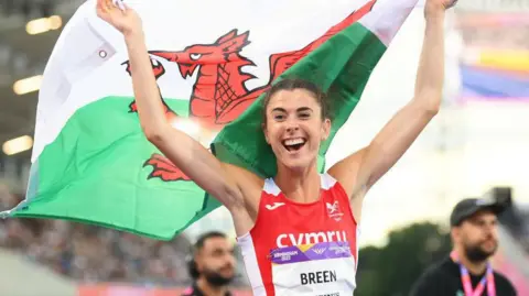 Olivia Breen holds the Welsh flag above her head after winning gold in the T38 100m at the Birmingham Commonwealth Games in 2022. 