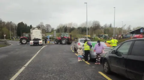 A road with trees in the background. The skies are grey. On the right is a row of cars, which appear to be at a standstill. There are two tractors and a lorry-cab parked blocking the road. Several people are stood around one of the cars. 
