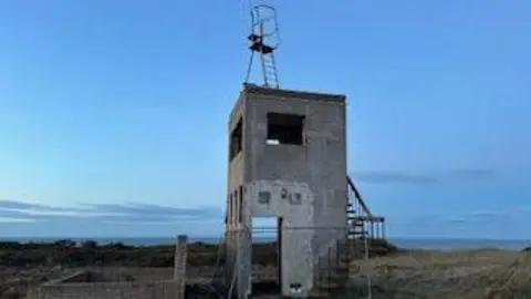 PHILIP CORKILL The existing lookout post, which is a run down light grey structure with stairs around it and a metal lookout point on top. It is in a rural area on a clear day.