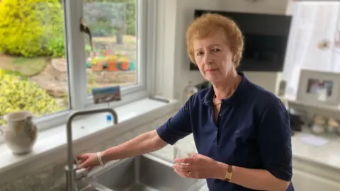 A woman stands in the kitchen next to the sink. She has one hand on the tap and in the other hand is holding a glass of water.  