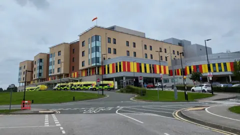 A hospital with ambulances parked outside it. The hospital building is several storeys high, and has red and yellow cladding on the lower part.