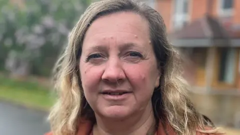 Lucy Nethsingha is standing on a residential street outside some homes, she is wearing a brown suede jacket.