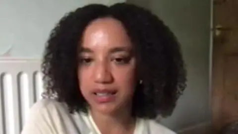 Close up of woman's face who is sitting in lounge near radiator. She has black curly hair and brown eyes.