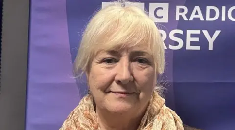 BBC Deputy Karen Wilson has blonde hair and is standing in front of a BBC Jersey purple banner. 
