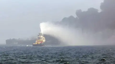 A tug boat firing its water jet at a burning tanker in the North Sea as smoke billows into the sky