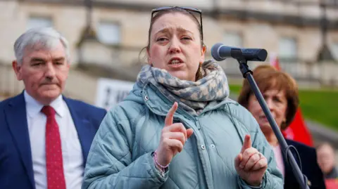 PA Media DUP MLA Deborah Erskine, standing in front of a microphone, wearing a light blue coat and a blue and cream scarf. There is a man standing behind her right shoulder. He has grey hair and is wearing a white shirt, blue suit and red tie. There is a woman behind her left shoulder, with short brown hair. She is wearing a pink top and a black suit. 