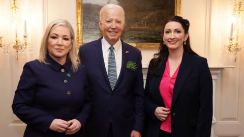 Michelle O'Neill and Emma Little-Pengelly standing either side of the US president, Joe Biden. Ms O'Neill looks serious while the other two are smiling.