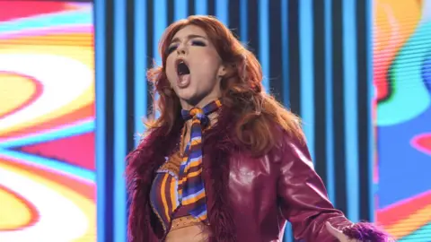 Jamie Hayter entering the arena for a wrestling match. She has long red hair, a maroon or purple jacket and ring gear that is purple, orange and red.