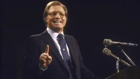 An old photo of Jimmy Swaggart shows him wearing a suit and glasses as he preaches on television