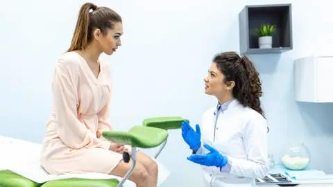 Stock photo of female gynecologist wearing blue medical gloves talks to a female patient wearing a peach coloured dress who is sat on a medical bed that has stirrups attached to the end