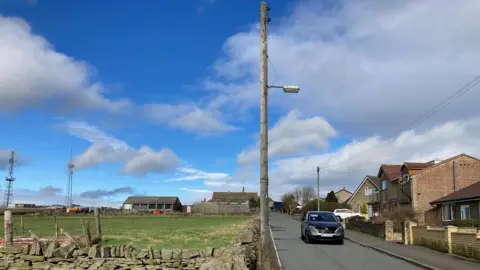 A green field with a dry stone wall and a grey road running to one side