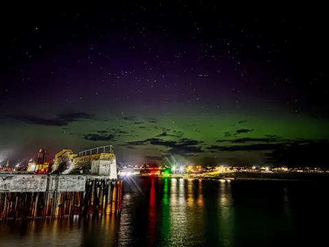 Paul Steven The Northern lights cast a green light over Wock harbour, while the bright lights of the town shine in the distance