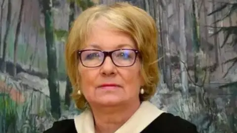 Marie Anderson is a middle-aged woman with blond hair and black-rimmed glasses. She is wearing a black jumper with white sleeves and neck and is at a desk with a a painting of trees in the background