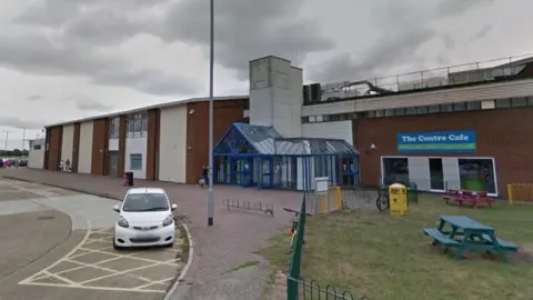A leisure centre building with a blue, glass front entrance and green space around it with a white car parked on the road in front of it 