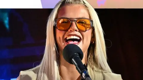 Skye Newman smiles behind a mic. She has long blonde hair and wears orange sunglasses