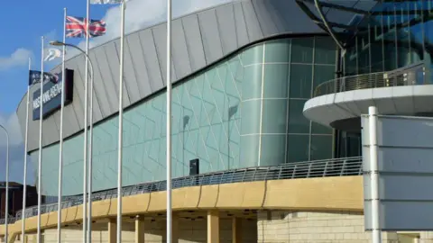 The exterior of the M&S Bank Arena with large glass opaque windows and a union flag and a black sign with white writing which reads M&S Bank Arena