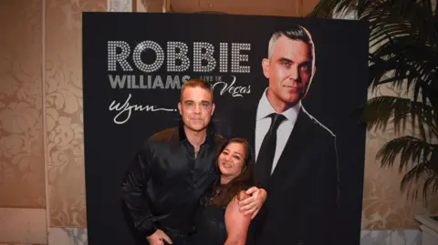 Rita Gulrajani A woman in a black dress stands next to Robbie Williams, who has his arm around her. They smile at the camera and stand in front of a black board with Robbie's face on it.