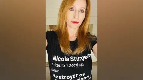 JK Rowling JK Rowling looking right at the camera. The picture is a selfie. She has long orange hair coming down below her neck. She has red lips and blue eyes. She is wearing a black T-shirt with white writing on it. The writing reads 'Nicola Sturgeon'. Below that is the word 'noun'. Below that are the words 'destroyer of women's rights'. However the last two words are obscured by the photo being cut off.