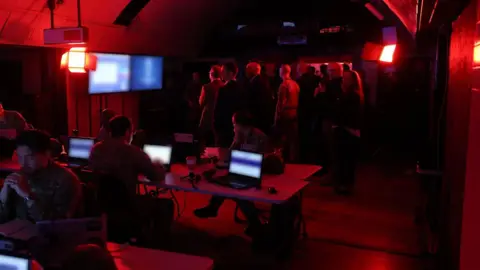 British Army A group of people stand in front of screens in a temporary command post - it is lit red. 