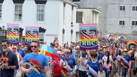 A large group of people participate in a Pride parade, holding rainbow flags and wearing colorful clothing. Several individuals carry signs with the RBC logo in rainbow colors. Buildings and a clear sky are visible in the background.