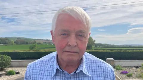 A man with silver hair and wearing a blue checked shirt, stands in a garden. In the background are green fields and trees.