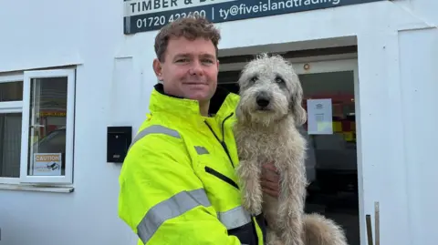 Tyrane Van Den Berg is smiling as he stands outside his office and is holding a dog in his arms. He has short blonde hair and blue eyes and is wearing a high-viz jacket. The light brown dog looks like a cockapoo as it has curly fur and it has ice blue eyes.