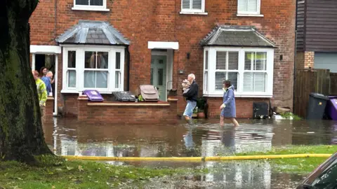 PA Media A couple walking through floodwater towards a home in Hitchin