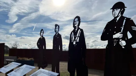 Sarah-May Buccieri/ BBC Four steel silhouette statues of women in military uniforms standing outdoors. In front of them are plaques displaying details about their contributions. The sky is blue with light clouds. 