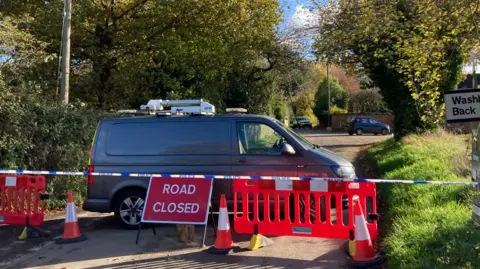 Luke Deal/BBC Part of Elm Lane in Copdock is closed off by blue and white police tape, a red 'road closed' sign, bright orange barriers and orange cones. A grey van is parked behind the cordon, it has been left across the road and would prevent any people or vehicles from getting past the road block. 