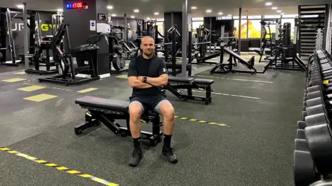 ROB WARD Gym manager Rob Ward sitting in his gym with yellow tape marking floor areas.