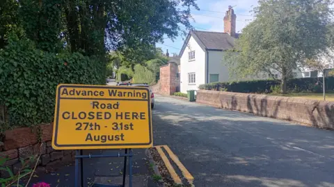 A yellow road sign warning of a road closure between 27-31 August