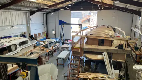 Owen Sennitt/BBC A boat workshop can be seen with two vessels currently being built or repaired, with wooden frames around them.