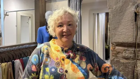 A woman with grey curly hair and a patterned jumper is standing in a room with stone walls and a large mirror