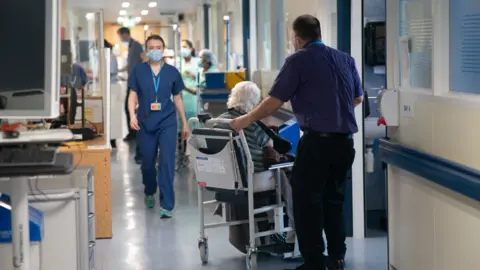 PA Media A stock image of a busy hospital. A doctor in blue scrubs walks towards the camera, while and elderly person is supported in a wheelchair. 