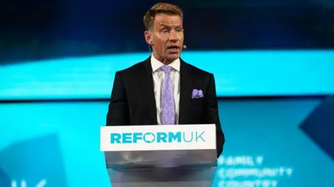 New chairman of Reform UK David Bull standing behind a podium with the Reform UK logo, speaking indoors against a plain backdrop
