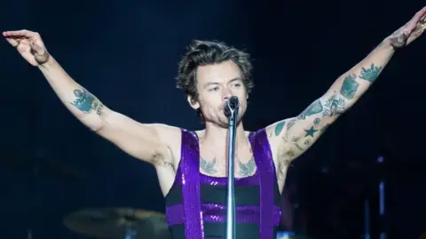 Harry Styles, who is wearing a sparkly purple and black top, has his arms in a V-shape in the air as he sings into a microphone. 