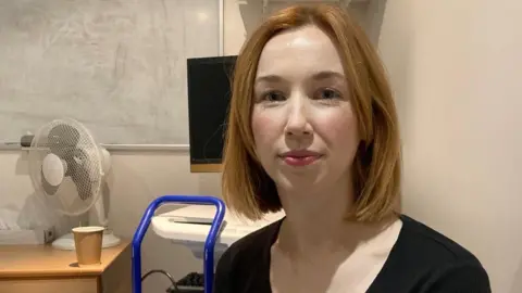 BBC Molly Macdonald has auburn hair and is wearing a black top. She is looking straight at the camera. A paper cup and a fan are on a desk behind her. 