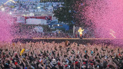 Getty Images A woman standing and singing on a catwalk surrounded by fans with ticker-tape filling the sky.