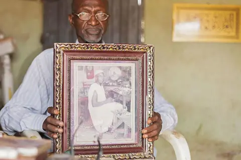 Ifiokabasi Ettang / BBC Bala Muhammad, mengenakan kacamata berbingkai kawat dan kemeja biru lengan panjang bergaris-garis, duduk di kursi kayu bercat putih sambil memegang foto hitam-putih berbingkai kayu lebar milik ayahnya, yang merupakan digambarkan sedang menatap kamera saat dia duduk di bangku bengkelnya. Dia mengenakan boubou tanpa lengan. Kipas meja terlihat di latar belakang dan jam pendulum tergantung di dinding