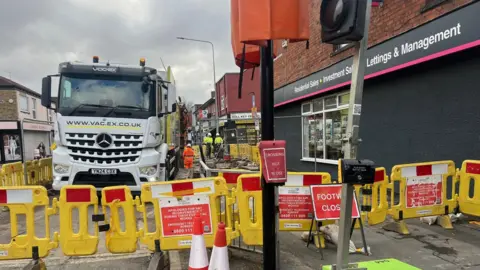 Image of roadworks. A large white lorry is parked on the left surrounded by yellow temporary barriers. Behind the lorry is a large hole in the ground, which is being dug. There are two orange cones in front of the yellow barriers and red signs reading: 'footpath closed'.